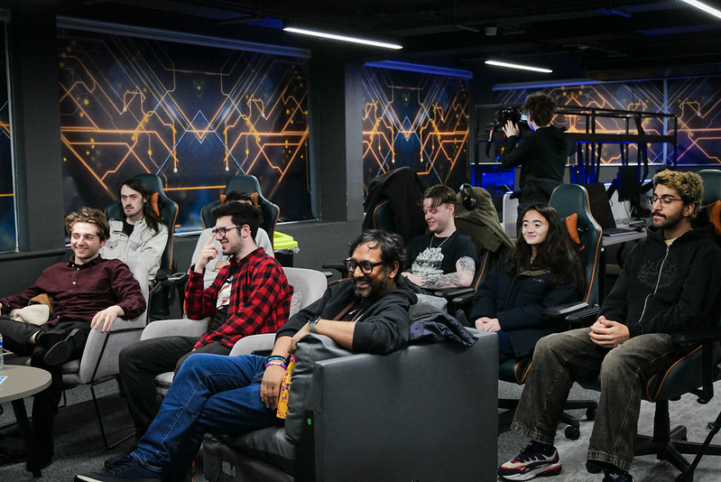 Image from a previous UWL games event where students and visitors are sat on sofas and gaming chairs while facing towards an unseen presenter. They are smiling, laughing, and reacting to something happening off-camera.