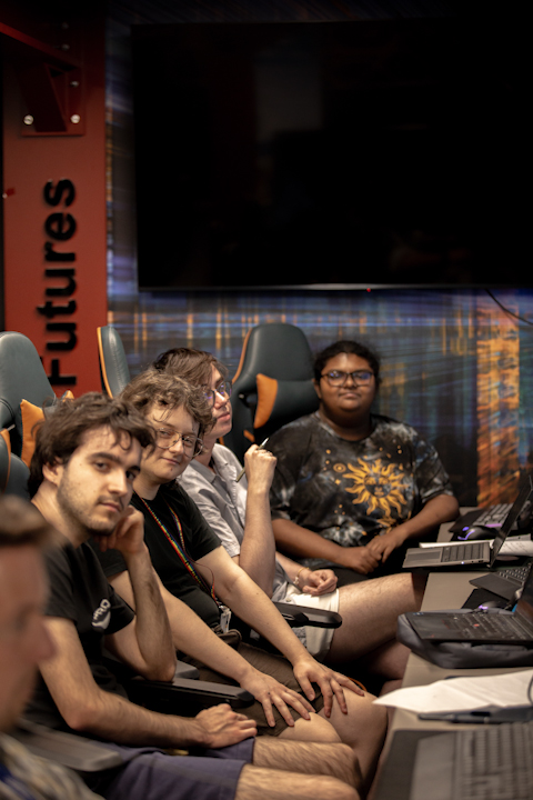 4 young developers, the team members of the team Microplastics, sat at a desk on gaming chairs who are facing the camera with neutral expressions